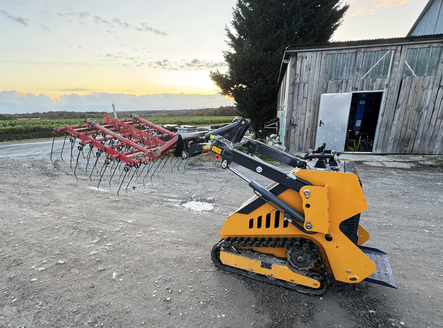Chargeur sur chenilles Zentrax 800 attelé à une herse devant un bâtiment agricole